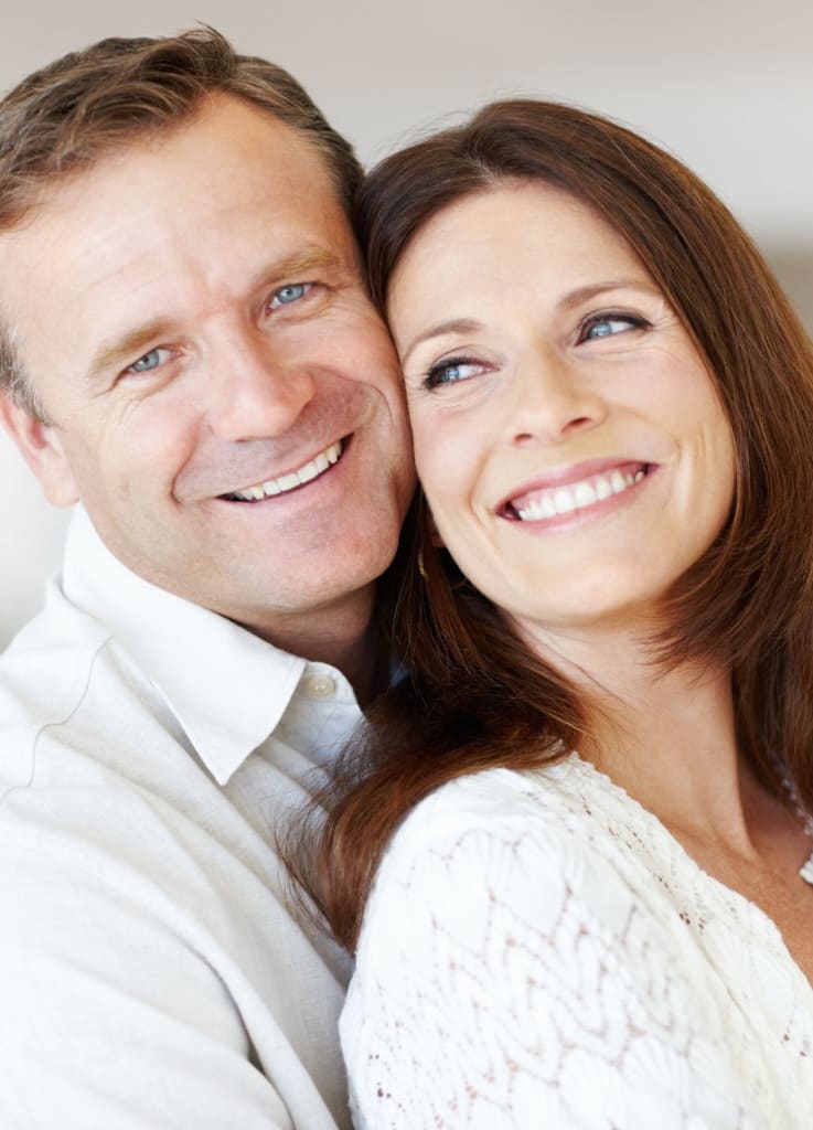 A smiling couple stands closely together, looking off camera. They are both wearing light-colored clothing, embodying the relaxed elegance one might feel after a visit with their concierge doctor.